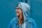 Eine junge Frau mit Brille, grauer Mütze und Jeansjacke lacht herzlich zur Seite. Sie steht vor einer blauen Wand. Das Bild vermittelt eine fröhliche, lockere und entspannte Stimmung mit einem natürlichen Lächeln.