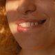 Detailaufnahme der unteren Gesichtshälfte einer Frau mit rotblondem, lockigem Haar. Sie lächelt zurückhaltend, wobei die oberen Zähne und sommersprossige Haut im warmen Sonnenlicht gut zu erkennen sind.