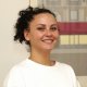 Freundliches Porträt von Angelica Amato, Dentalassistentin in Ausbildung der Zahnarztpraxis & Oralchirurgie Spitalgasse in Bern. Lächelnde junge Frau mit dunklen, hochgesteckten Locken und weisser Berufskleidung vor hellem Hintergrund.