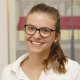 Porträtbild von Jeannine Fleischlin, Dentalhygienikerin HF (Swiss Dental Hygienists), mit Brille, zusammengebundenen Haaren und weissem Poloshirt, lächelnd vor einem unscharfen Hintergrund mit Berner Altstadtfassade.