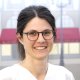 Portraitfoto von Dr. Daniela Zeller, Fachzahnärztin für rekonstruktive Zahnmedizin, mit Brille und freundlichem Lächeln vor unscharfem Berner Hintergrund mit roter Markise. Sie trägt eine helle Berufskleidung.